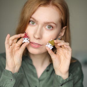 Pu&ograve; includere: Due piccole figure di funghi all'uncinetto. Un fungo ha un cappello rosso a pois bianchi, l'altro un cappello verde. Entrambi hanno corpi bianchi e facce sorridenti. Le figure sono tenute davanti a una persona.