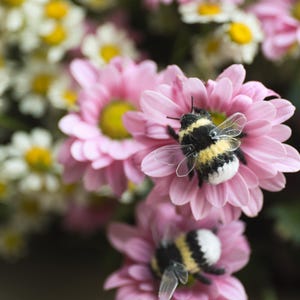 May include: Close-up of pink and white flowers with yellow centers. Two handmade bumblebees with black, yellow, and white bodies and clear wings are perched on the flowers. Other flowers are in the background.