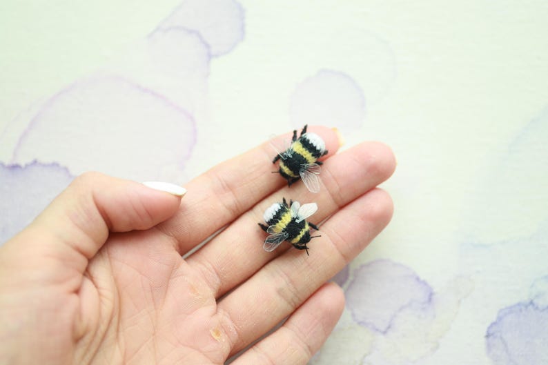 May include: Two miniature bumblebee figurines with black, yellow, and white details. The bees have transparent wings and are resting on a hand. The background is a light, watercolor-style wash with purple hues.