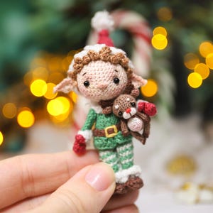 May include: A handmade crochet elf doll wearing a green and white outfit with a red hat and mittens. The elf is holding a small brown reindeer. The doll has a tan face with brown hair and is wearing a brown belt with a gold buckle. The background is blurred with Christmas decorations.
