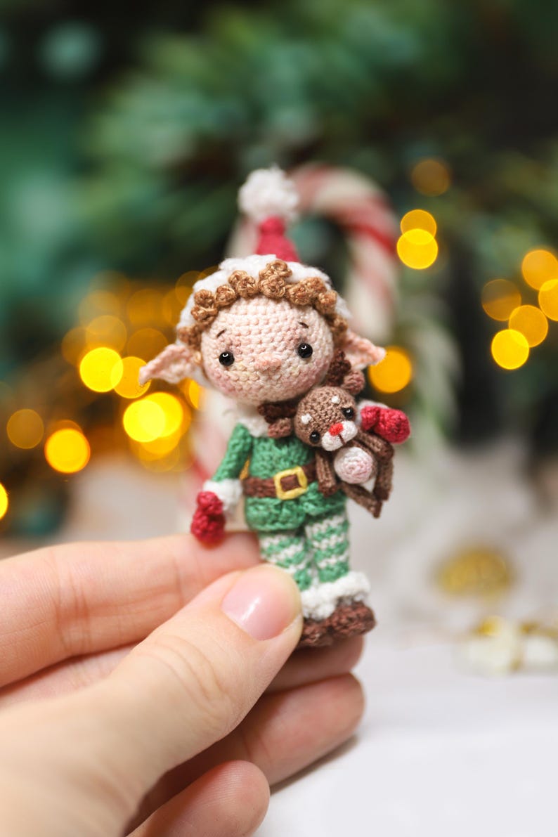 May include: A handmade crochet elf doll wearing a green and white outfit with a red hat and mittens. The elf is holding a small brown reindeer. The doll has a tan face with brown hair and is wearing a brown belt with a gold buckle. The background is blurred with Christmas decorations.