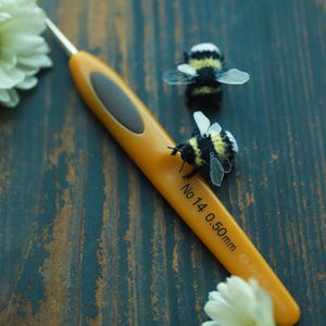 May include: An orange crochet hook with a black grip and the text "No 14 0.50mm" is next to two small, handmade bee figurines with yellow and black stripes and clear wings. White flowers are in the background.
