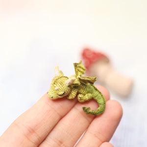 Tiny green micro crochet dragon resting on fingers, handmade amigurumi miniature with curled tail, small wings and horns, sleeping fantasy creature made with fine thread, scale shown by human hand.