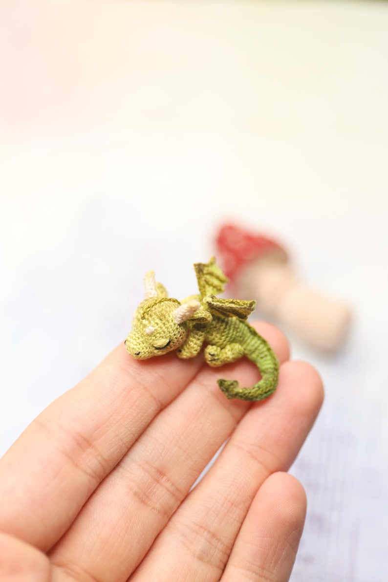 Tiny green micro crochet dragon resting on fingers, handmade amigurumi miniature with curled tail, small wings and horns, sleeping fantasy creature made with fine thread, scale shown by human hand.