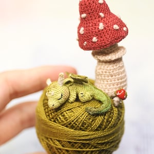 May include: A handmade crochet sculpture featuring a sleeping green dragon atop a ball of olive green yarn. A red mushroom with white spots and a small red strawberry are also included. The sculpture is held in a hand.