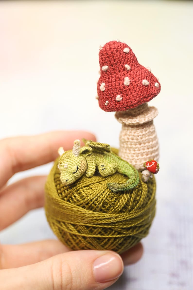 May include: A handmade crochet sculpture featuring a sleeping green dragon atop a ball of olive green yarn. A red mushroom with white spots and a small red strawberry are also included. The sculpture is held in a hand.