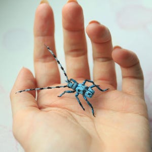 May include: A detailed, handmade insect sculpture in blue and black, with long antennae, resting on a hand. The insect's body and legs are textured. The background is a soft, watercolour-like wash of colour.
