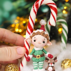 May include: A crocheted Christmas elf doll with a green outfit, red gloves, and a brown belt stands next to a candy cane. A small crocheted reindeer and gold ornaments are also present, creating a festive holiday scene.
