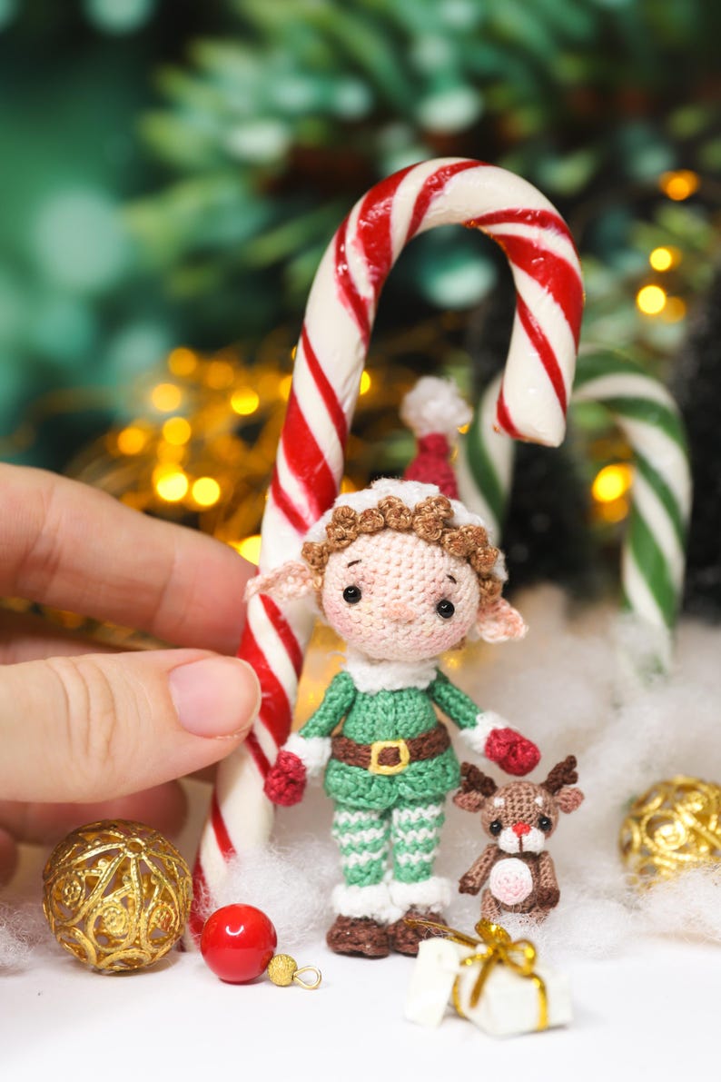 May include: A crocheted Christmas elf doll with a green outfit, red gloves, and a brown belt stands next to a candy cane. A small crocheted reindeer and gold ornaments are also present, creating a festive holiday scene.