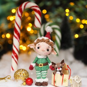 May include: A crocheted Christmas elf doll with a green outfit, red gloves, and a white collar stands next to a small reindeer in a gift box. A candy cane and Christmas ornaments are in the background, with a blurred bokeh effect.