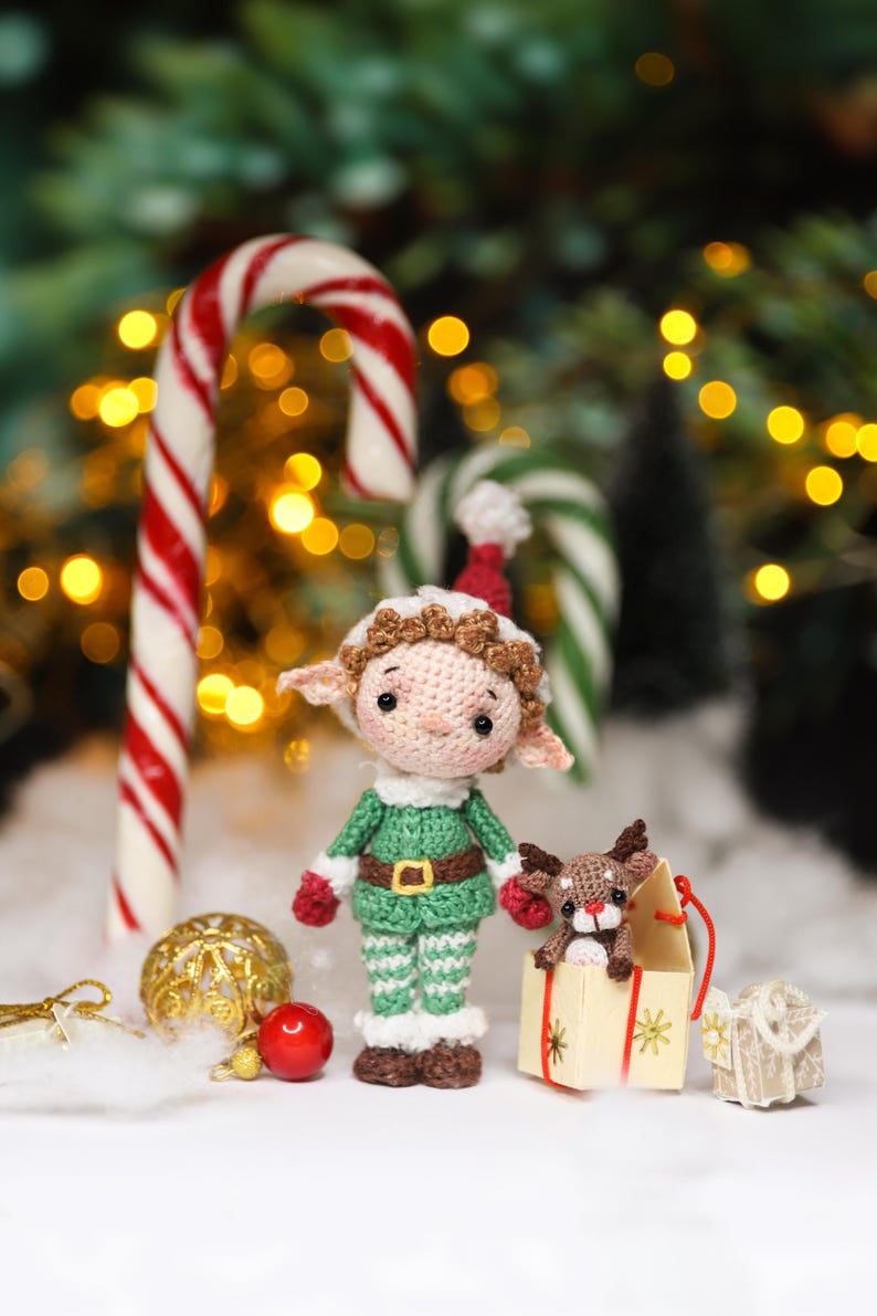 May include: A crocheted Christmas elf doll with a green outfit, red gloves, and a white collar stands next to a small reindeer in a gift box. A candy cane and Christmas ornaments are in the background, with a blurred bokeh effect.
