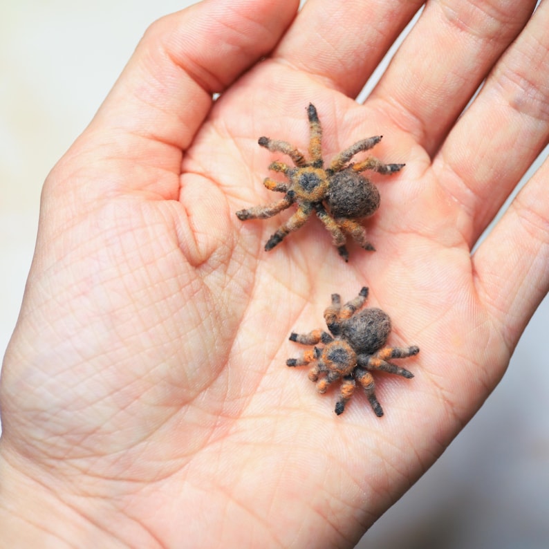 May include: Two small, detailed spider figurines with orange and black markings are displayed in a person's open palm. The spiders have a fuzzy texture and realistic features, suggesting they are handmade crafts or toys.