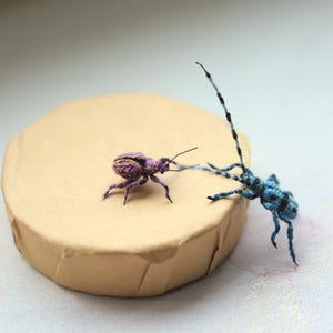 May include: Two handmade insect sculptures, one purple and one blue, are positioned on a tan, round platform. The purple insect has a striped pattern, while the blue insect has long antennae. The background is a soft, blurred white.