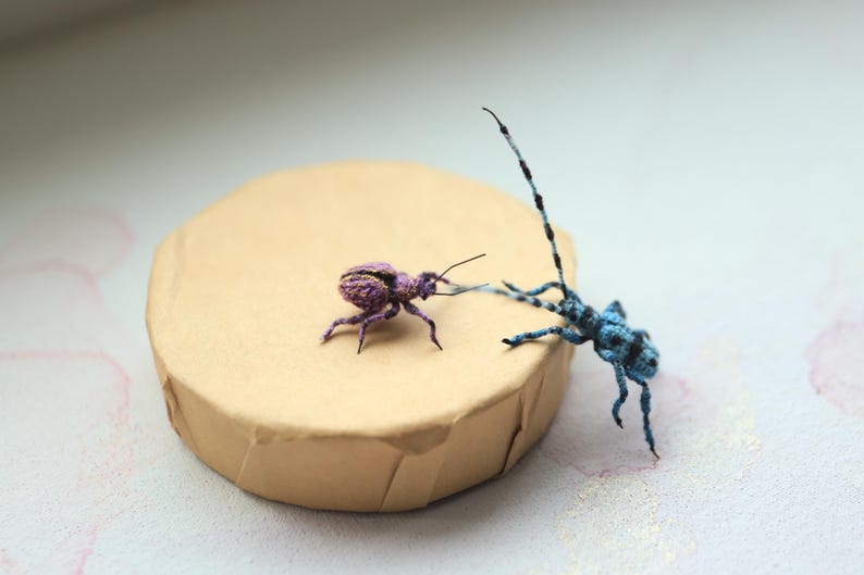 May include: Two handmade insect sculptures, one purple and one blue, are positioned on a tan, round platform. The purple insect has a striped pattern, while the blue insect has long antennae. The background is a soft, blurred white.