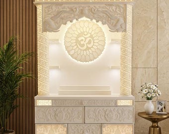 May include: Ornate white cabinet with intricate carvings, illuminated by soft lighting. Features a central Om symbol and Sanskrit text. Includes a small side table with a vase of flowers and a framed photo. A decorative piece for home decor.