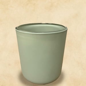 May include: A light green, cylindrical glass tumbler with a rounded base. The rim of the glass has a darker green accent. The glass is opaque and sits on a light brown background.