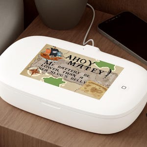 Può includere: Un dispositivo rettangolare bianco con uno schermo che mostra una mappa e il testo "Ahoy Matey! Me Battery Be Lower Than A Sea Slug's Belly." Una piccola pianta in vaso, uno smartphone e occhiali sono su un comodino di legno.