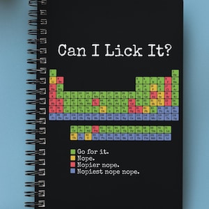 May include: Black spiral-bound notebook with the text "Can I Lick It?" and a periodic table design. The notebook also includes the phrases "Go for it," "Nope," "Nopier nope," and "Nopiest nope nope." A pencil, glasses, and headphones are also visible.