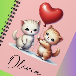 May include: A spiral-bound notebook with a pink cover featuring two cartoon kittens and a red heart-shaped balloon. The word "Olivia" is written in cursive at the bottom. The notebook has a green spine.
