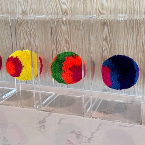 May include: A display of six colorful pom-poms, each suspended within a clear acrylic box. The pom-poms vary in color, including combinations of gray, blue, red, yellow, and green. The boxes are arranged in a row against a light wood-grain background.