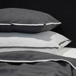 Pode incluir: Uma pilha de roupa de cama cinza e branca com uma borda branca. A roupa de cama é feita de um tecido macio e confortável e é perfeita para uma noite de sono aconchegante.