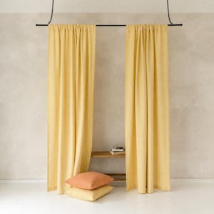 May include: Two floor-length yellow curtains hang from a black metal rod suspended by chains. Below the curtains, two square throw pillows are stacked on top of each other, with the top pillow in a muted orange and the bottom one in yellow.