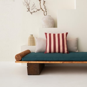 May include: A modern wooden daybed with a teal cushion and two cylindrical wooden armrests. It features a striped red and pink pillow and two neutral-colored pillows. The daybed is supported by two large wooden blocks.