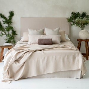 Peut inclure: Un lit en lin beige avec un coussin de soutien-gorge brun et deux oreillers blancs. Le lit a une tête de lit beige et repose sur un sol blanc. Il y a deux plantes en pot de chaque côté du lit.