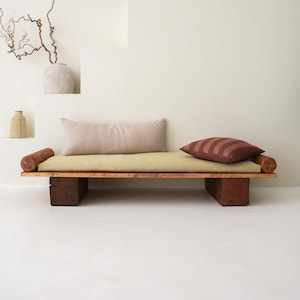May include: A contemporary wooden daybed featuring a light green cushion, two cylindrical wooden armrests, and two pillows. One pillow is light pink, the other has brown stripes. The daybed rests on two dark brown wooden blocks.