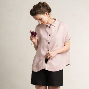 May include: A pink linen shirt with short sleeves and a button-up front. The shirt is slightly oversized and has a relaxed fit. The model is wearing black linen shorts.