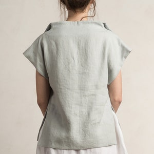 May include: A light blue linen top with a wide, folded neckline and short sleeves. The top has a relaxed fit and a slightly longer back hem.