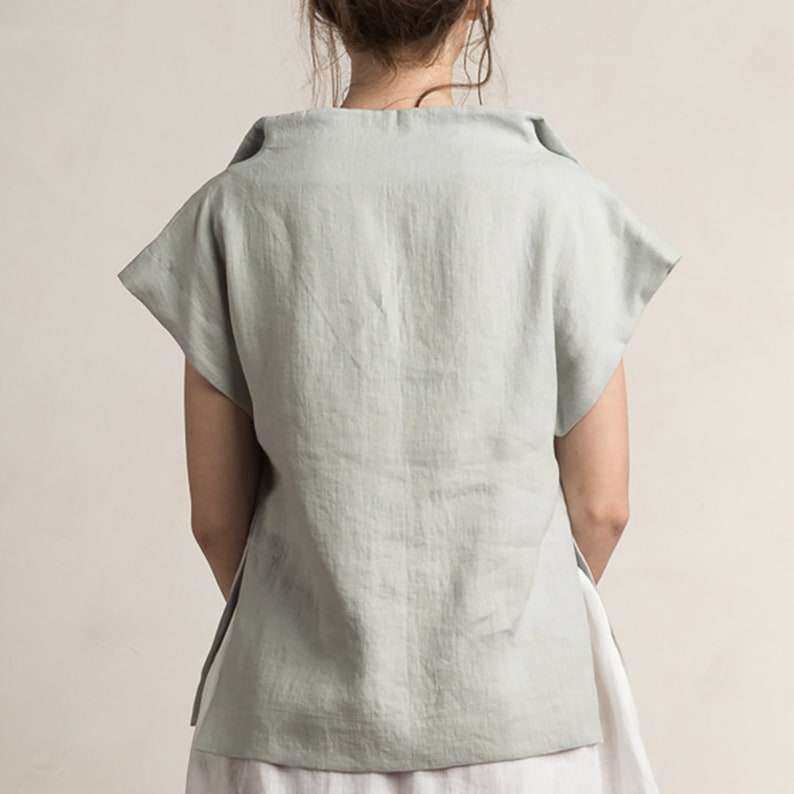 May include: A light blue linen top with a wide, folded neckline and short sleeves. The top has a relaxed fit and a slightly longer back hem.