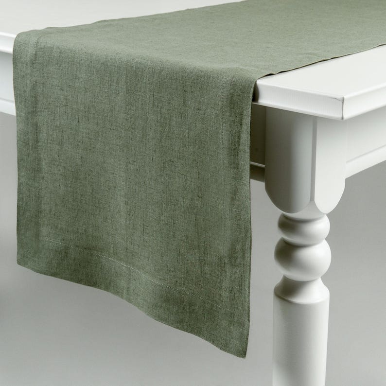 May include: A sage green linen table runner draped over a white table. The runner has a textured appearance and a finished hem. The table has a classic design with a rounded leg.