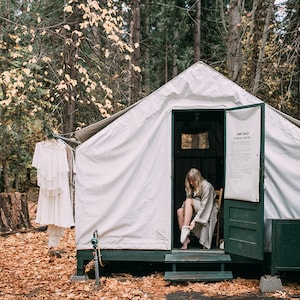 May include: A woman in a white robe sits on a wooden chair inside a white canvas tent with a green door. The tent has a sign that reads "Camp Rules".