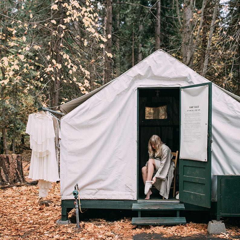 May include: A woman in a white robe sits on a wooden chair inside a white canvas tent with a green door. The tent has a sign that reads "Camp Rules".