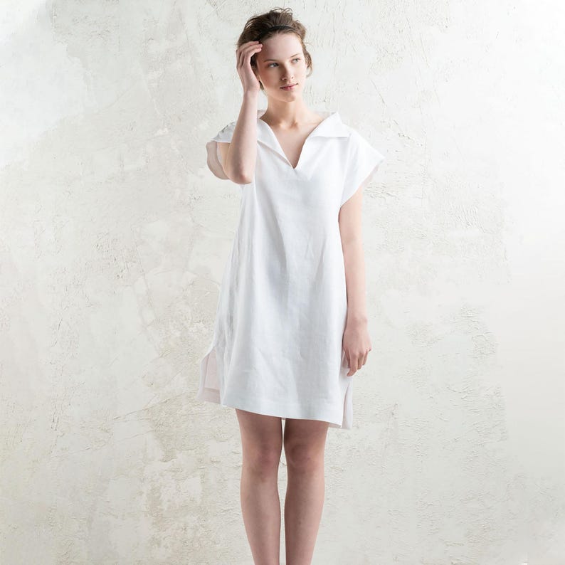 May include: A white linen dress with a V-neck and short sleeves. The dress has a relaxed fit and a slightly longer length in the back.