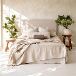Puede incluir: Una cama beige con cabecero y ropa de cama a juego. La cama está adornada con varias almohadas en diferentes tonos de beige y un pequeño cojín cilíndrico marrón. Dos pequeños taburetes de madera flanquean la cama, cada uno con una planta en maceta.