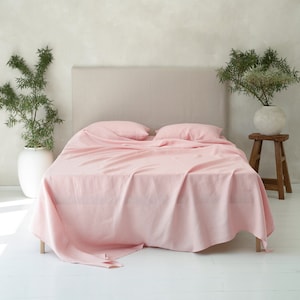 May include: A bed with a light pink linen sheet set. The bed has a neutral-colored headboard and two pillows. The room is decorated with plants in white vases and a wooden stool.