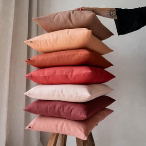 May include: A stack of eight decorative throw pillows in various shades of red, orange, and pink. The pillows are made of a soft, linen-like fabric and are arranged in a graduated colour scheme.