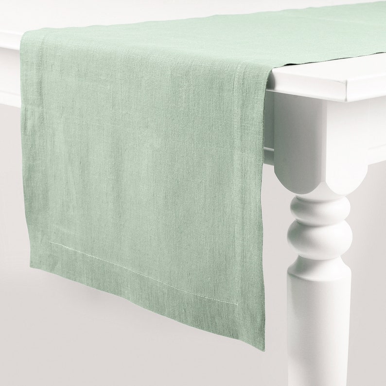 Peut inclure: Chemin de table en lin vert clair pos&eacute; sur une table blanche. Le tissu pr&eacute;sente une texture visible et un ourlet cousu. La table, avec son pied tourn&eacute;, apporte une touche d'&eacute;l&eacute;gance &agrave; l'ensemble.