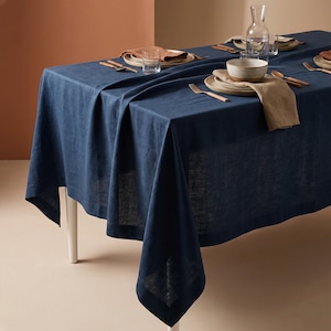 May include: A dark blue linen tablecloth draped over a rectangular table, set with ceramic plates, beige linen napkins, clear glass tumblers, a glass carafe, and metallic cutlery arranged for a formal dining experience.