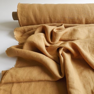 May include: A roll of tan-colored linen fabric with a raw edge. The fabric is draped and folded, showing its texture and color variations. The linen is a warm, earthy tone, suitable for various sewing and crafting projects.