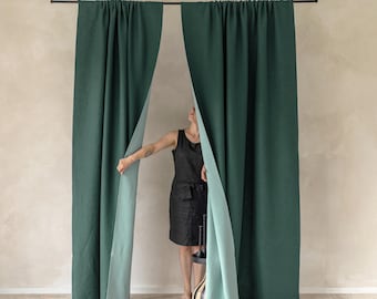 May include: Dark green linen curtains with a light green backing are suspended from a black rod. The curtains are partially open, revealing a person standing behind them. The curtains are hanging in front of a light beige wall.