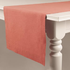 Puede incluir: Un camino de mesa de lino color coral drapeado sobre una mesa blanca. El camino de mesa tiene forma rectangular con un sutil detalle de borde. La mesa tiene un diseño de pata decorativa.