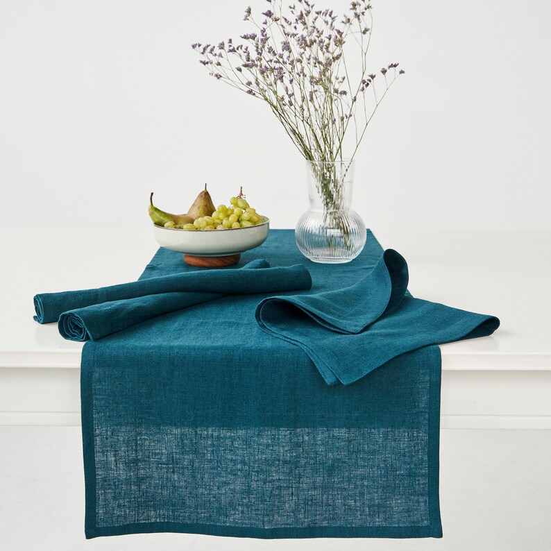 May include: A teal linen table runner with a subtle texture. The runner is folded in half and placed on a white table. A bowl of green grapes and pears sits on the table in front of a vase of purple flowers.