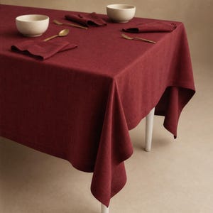 Op de afbeelding: Een dieprood tafelkleed gedrapeerd over een tafel, met twee gebroken witte kommen, twee gevouwen rode servetten en twee goudkleurige lepels. De stof heeft een zichtbare textuur en de tafel staat op witte poten voor een formele tafelsetting.