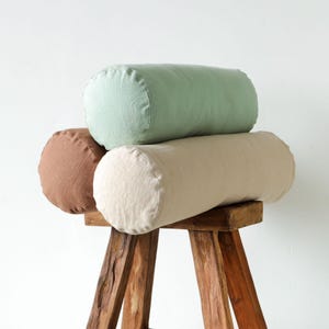 May include: Three cylindrical pillows in shades of brown, green, and beige are stacked on a rustic wooden stool. The pillows have a soft, textured appearance, suggesting a linen or cotton fabric. The stool has a natural wood finish.