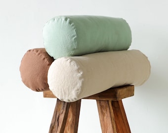 May include: Three cylindrical pillows in shades of brown, green, and beige are stacked on a rustic wooden stool. The pillows have a soft, textured appearance, suggesting a linen or cotton fabric. The stool has a natural wood finish.