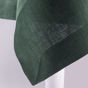 May include: A dark forest green fabric with a visible woven texture and a mitered corner hem. The material features a consistent deep green hue throughout, showcasing the interlaced threads of the textile weave and a clean, straight stitched border.