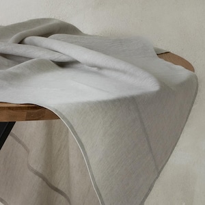 May include: A light gray linen tablecloth draped over a wooden table. The fabric has a subtle texture and a soft, flowing appearance. The tablecloth's edge is trimmed with a thin, lighter-colored border, adding a touch of elegance to the simple design.